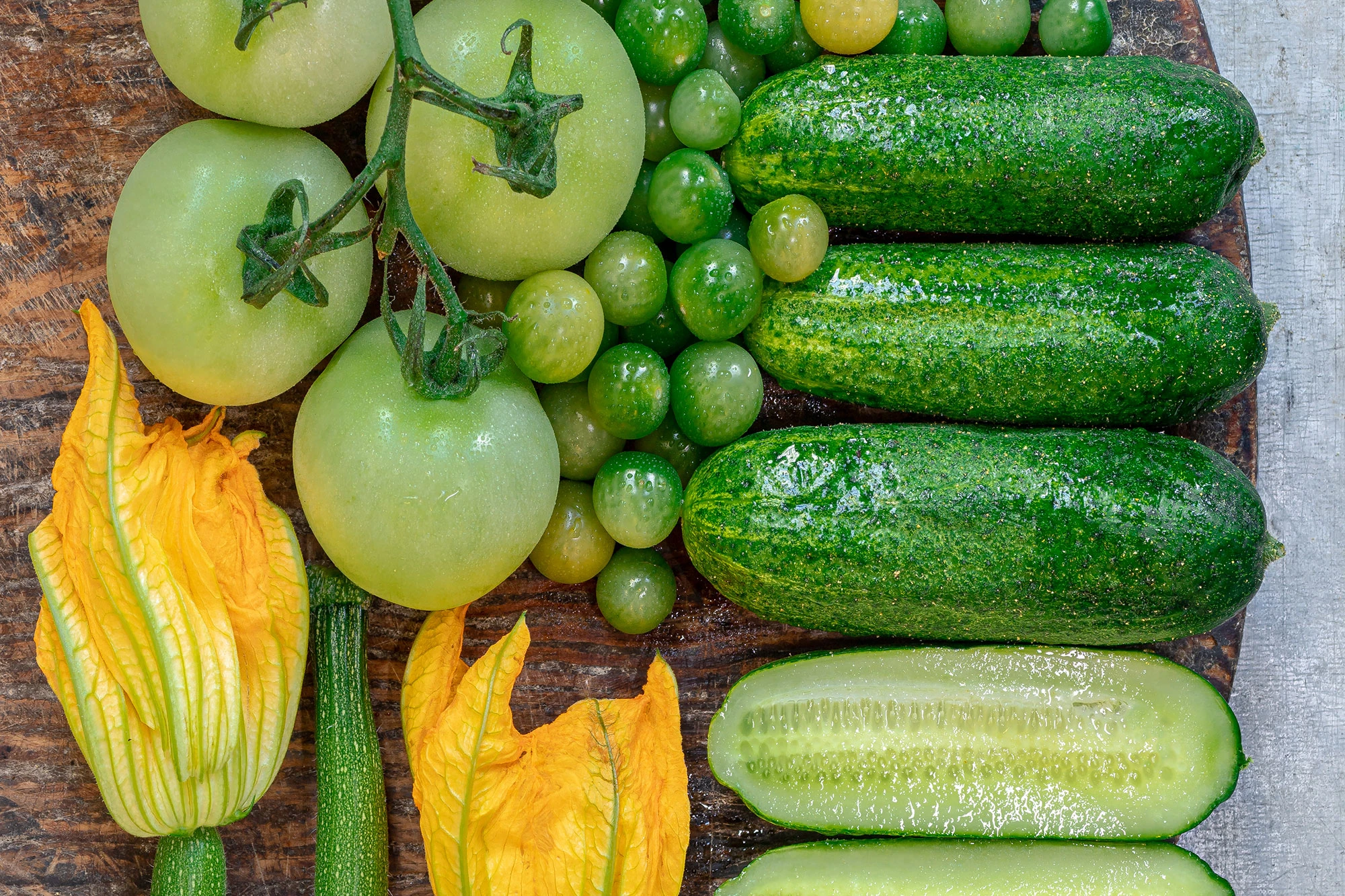 Sommargrönsaker som gröna tomater, gurka och zucchiniblommor på en träskärdbräda
